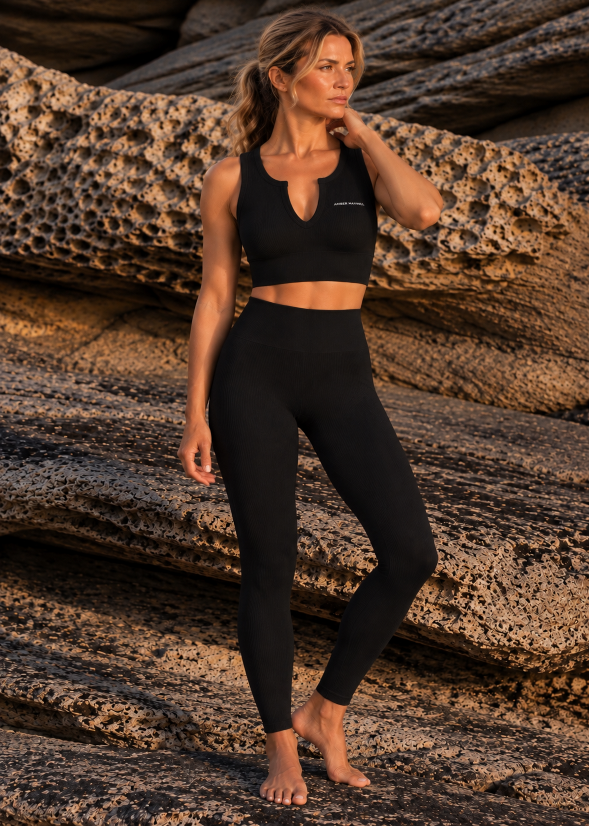 A woman modeling a black athletic crop top and matching high-waisted leggings, standing barefoot on textured rock formations outdoors.