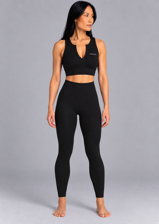 A woman modeling a black activewear set consisting of a sleeveless crop top with a keyhole neckline and matching high-waisted leggings, photographed against a solid gray studio background.
