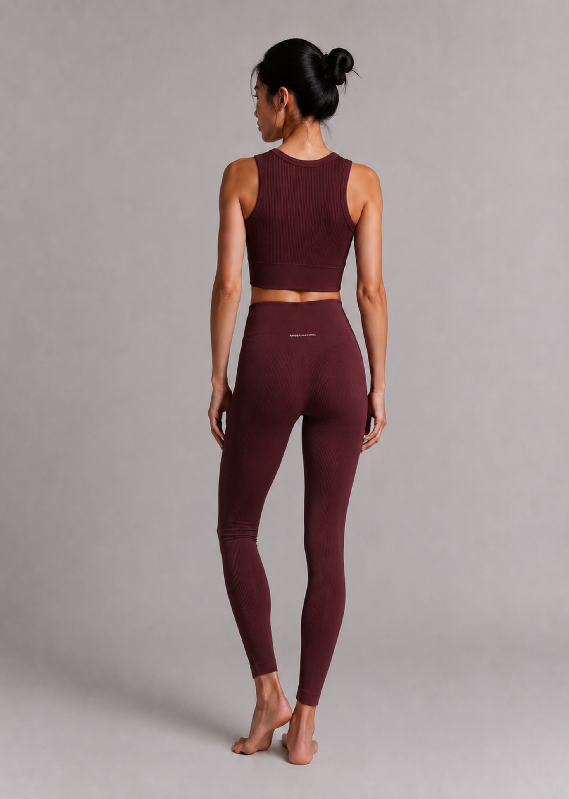 A woman is shown from behind wearing a matching burgundy activewear set consisting of a sleeveless crop top and high-waisted leggings, set against a plain gray studio background.
