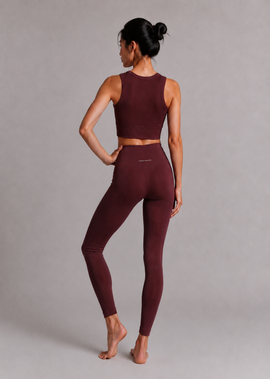 A woman modeling a matching burgundy activewear set consisting of a sleeveless crop top and high-waisted leggings, shown from a rear three-quarter angle against a neutral gray studio background.