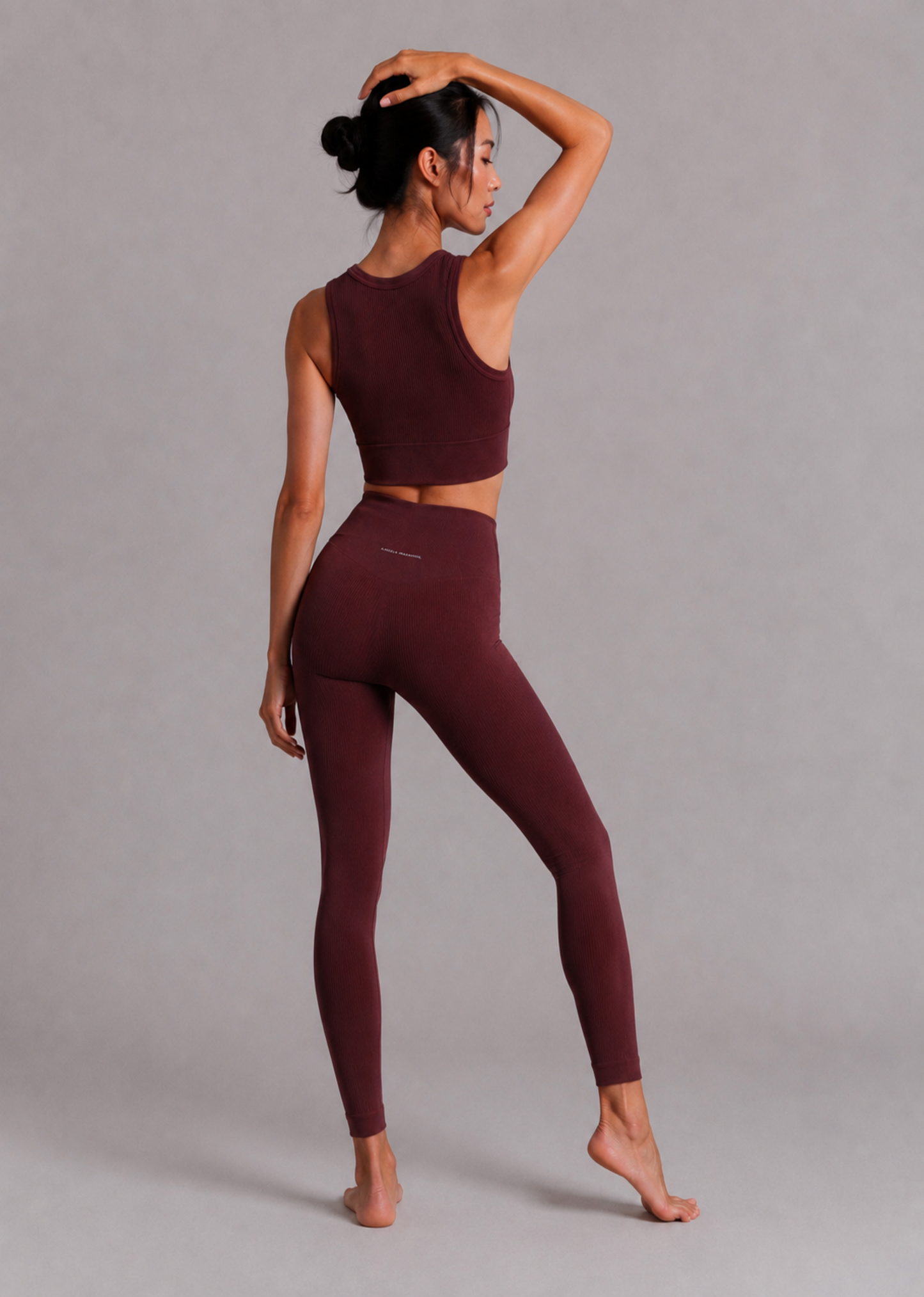 A woman modeling a matching burgundy activewear set consisting of a sleeveless crop top and high-waisted leggings, shown from a side-back angle against a neutral gray studio background.