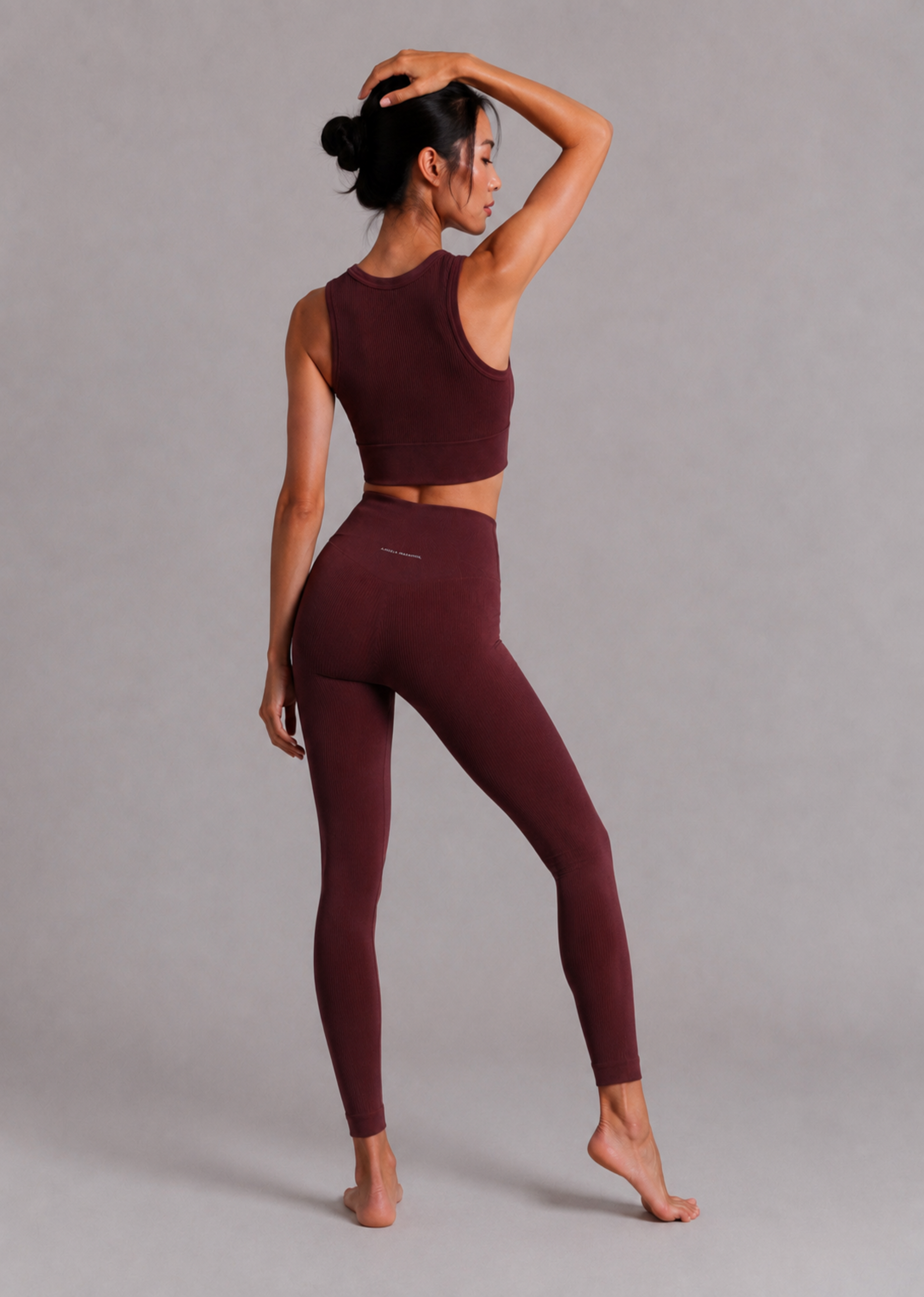 A woman modeling a matching burgundy activewear set consisting of a sleeveless crop top and high-waisted leggings, shown from a side-back angle against a neutral gray studio background.