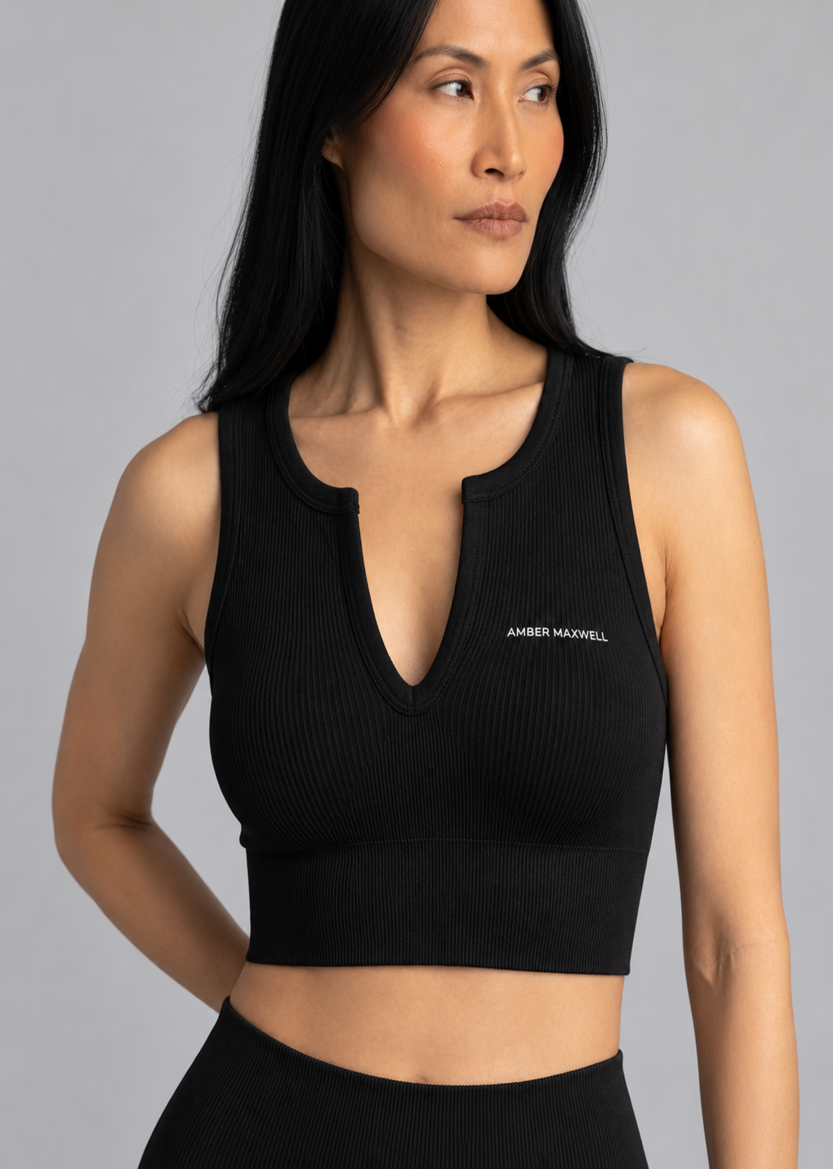 A woman modeling a black ribbed knit sports bra with a split neckline and the brand name 'AMBER MAXWELL' printed in white on the chest, set against a solid gray studio background.