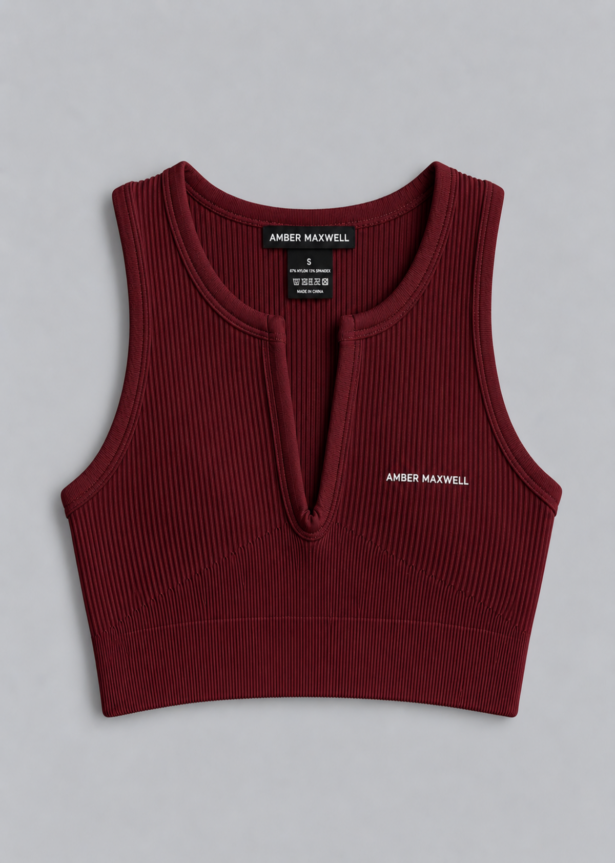 A sleeveless ribbed knit crop top in deep burgundy, featuring a split V-neckline with a small center tab, a black neck label reading 'AMBER MAXWELL', and white 'AMBER MAXWELL' text on the left chest, laid flat on a light gray background.
