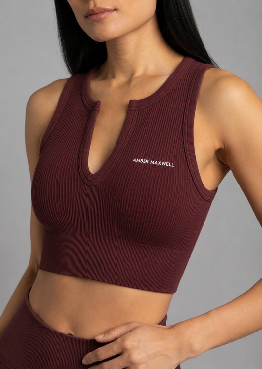 A close-up of a woman wearing a ribbed sleeveless crop top in deep burgundy. The top features a split neckline with a small keyhole cutout and the brand name 'AMBER MAXWELL' printed in white on the left chest.