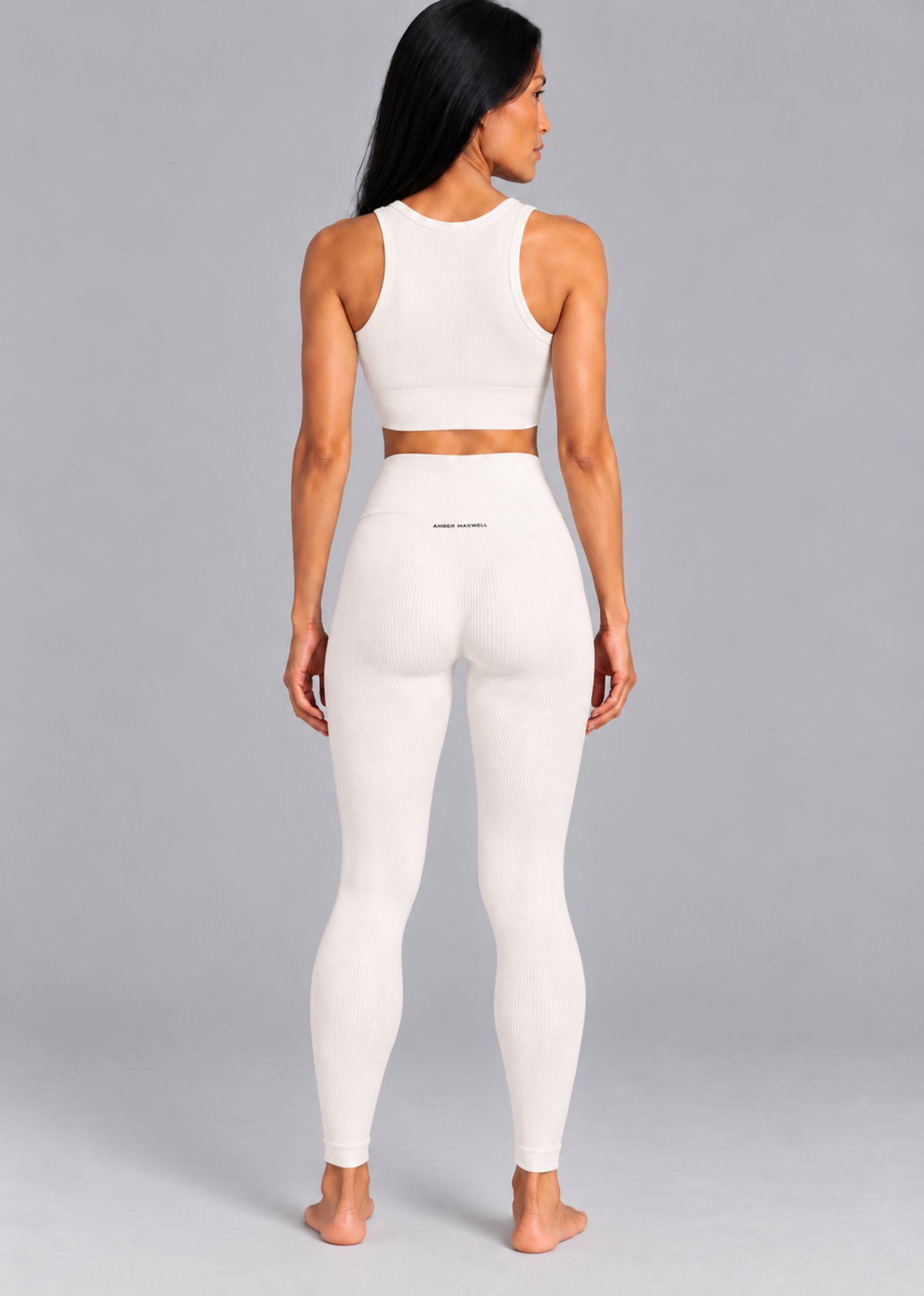 A woman is shown from the back wearing a matching white activewear set consisting of a sleeveless crop top and high-waisted leggings, set against a solid gray studio background.