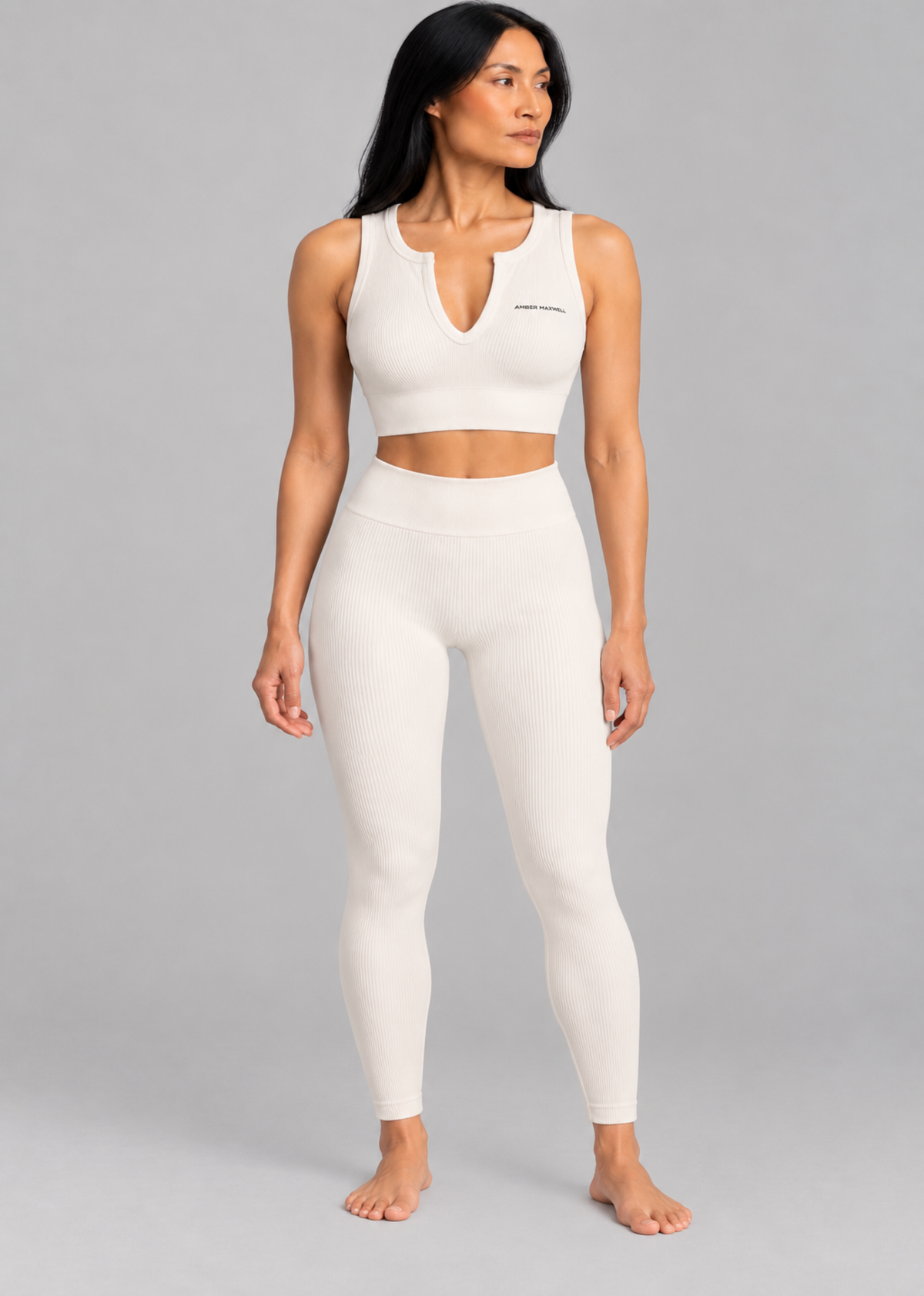 A woman modeling a matching off-white activewear set consisting of a sleeveless crop top with a keyhole neckline and high-waisted leggings, set against a solid gray studio background.