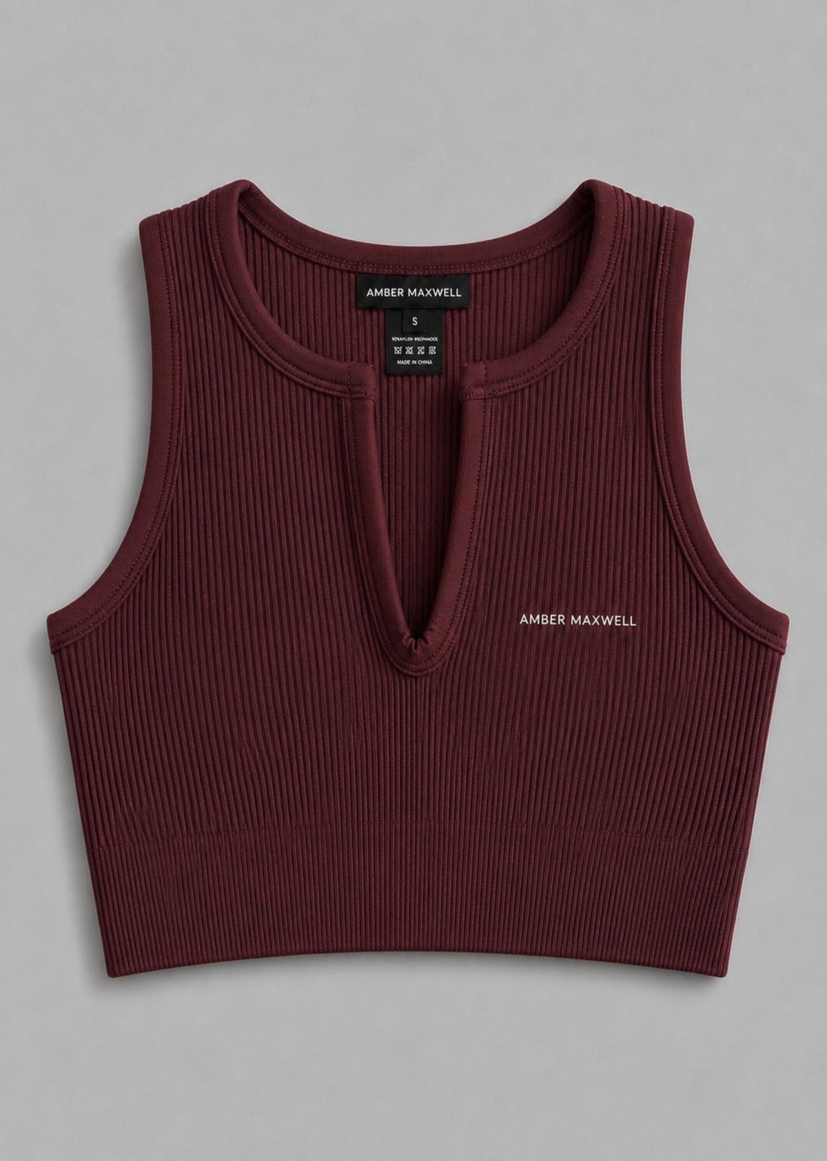 A ribbed knit sleeveless crop top in deep burgundy, featuring a split V-neckline and small white 'AMBER MAXWELL' text on the left chest, displayed flat against a neutral gray background.