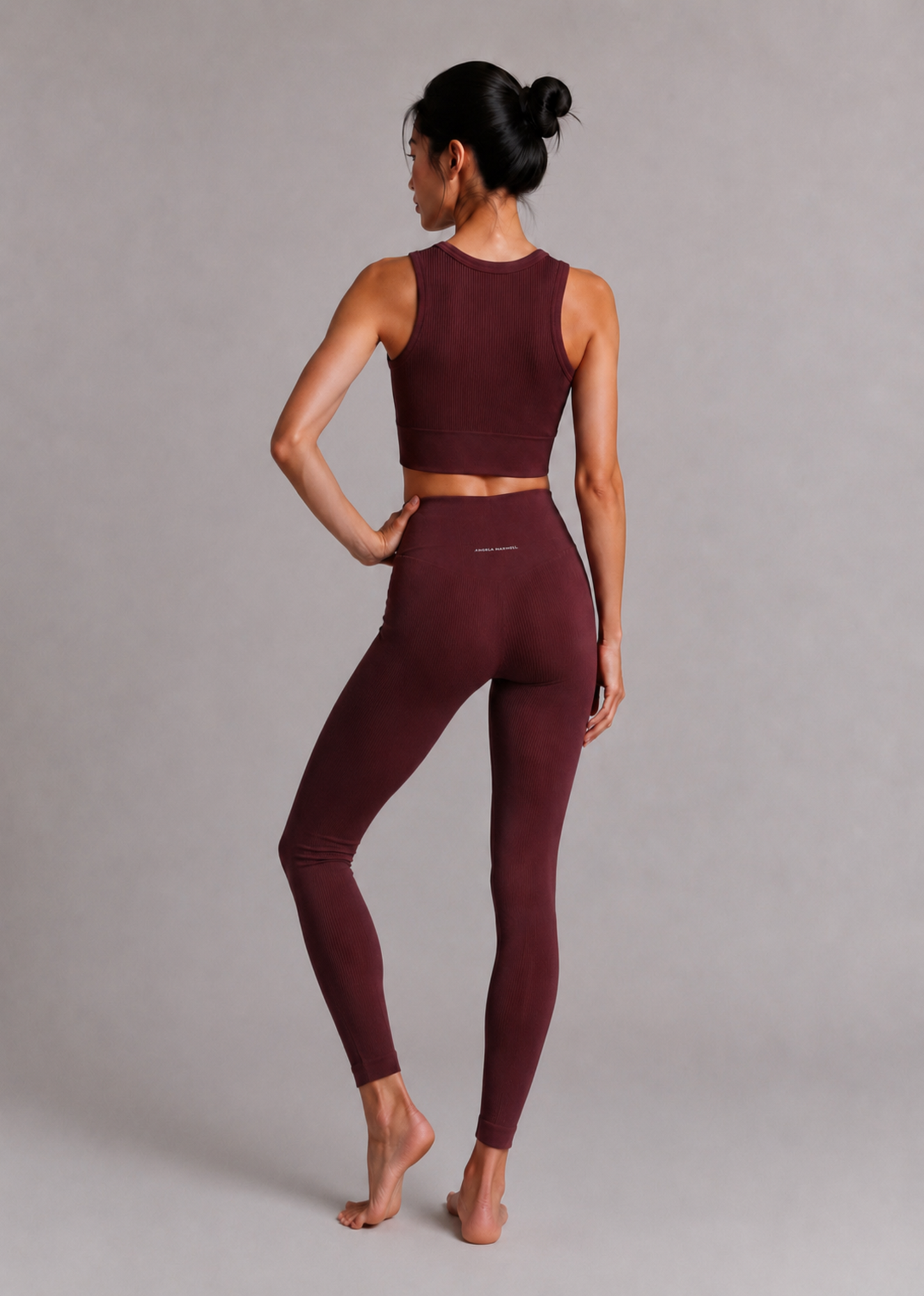 A woman modeling a matching burgundy activewear set consisting of a sleeveless crop top and high-waisted leggings, shown from a rear three-quarter angle against a neutral gray studio background.