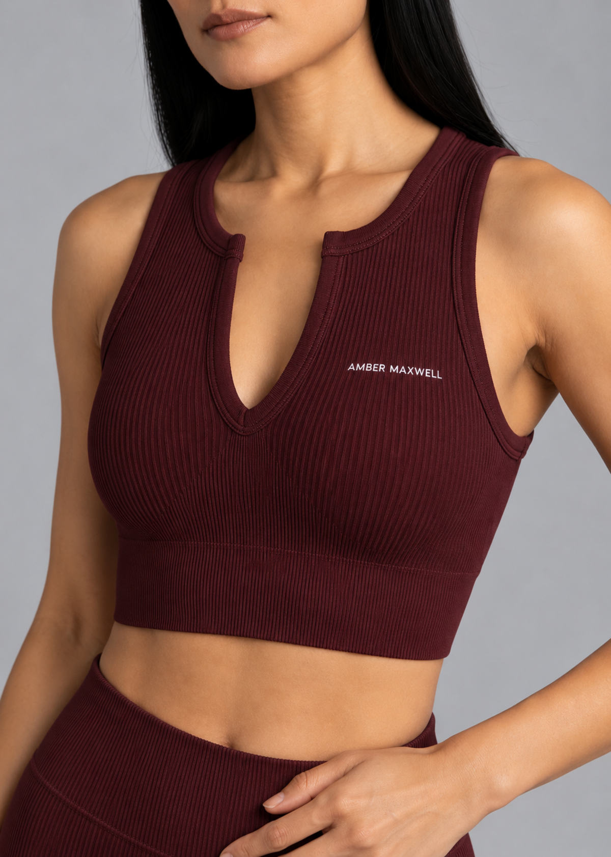 A close-up of a woman wearing a ribbed sleeveless crop top in deep burgundy. The top features a split neckline with a small keyhole cutout and the brand name 'AMBER MAXWELL' printed in white on the left chest.