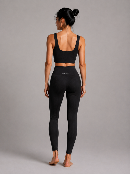 A woman stands barefoot against a plain gray studio background, photographed from the back. She is wearing a black ribbed sports bra and matching black high-waisted leggings with small white text on the back waistband.