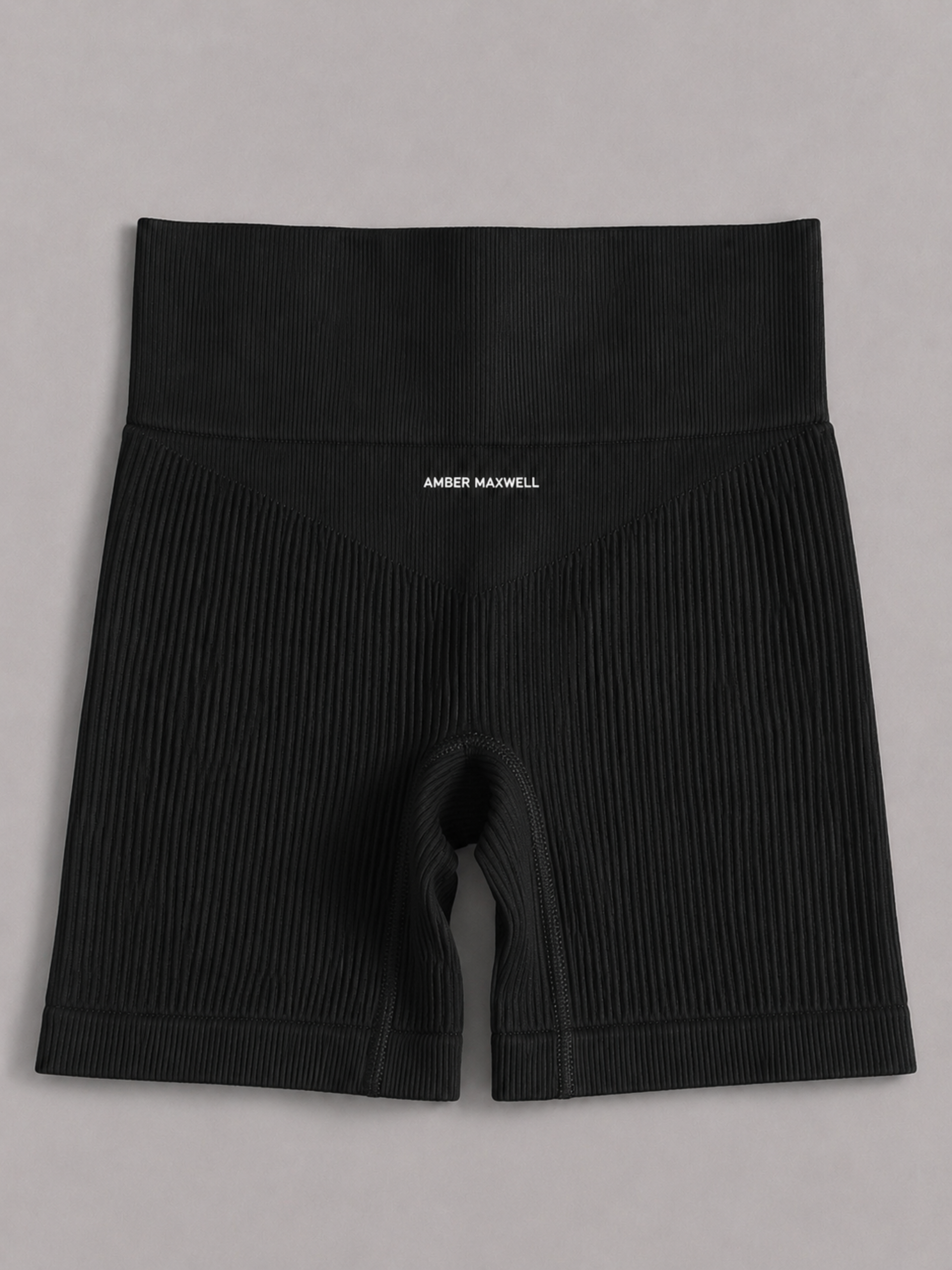 A ribbed knit sports bra in a black color, featuring a small white embroidered logo on the left chest that reads 'AMBER MAXWELL'. Gigi Shorts.