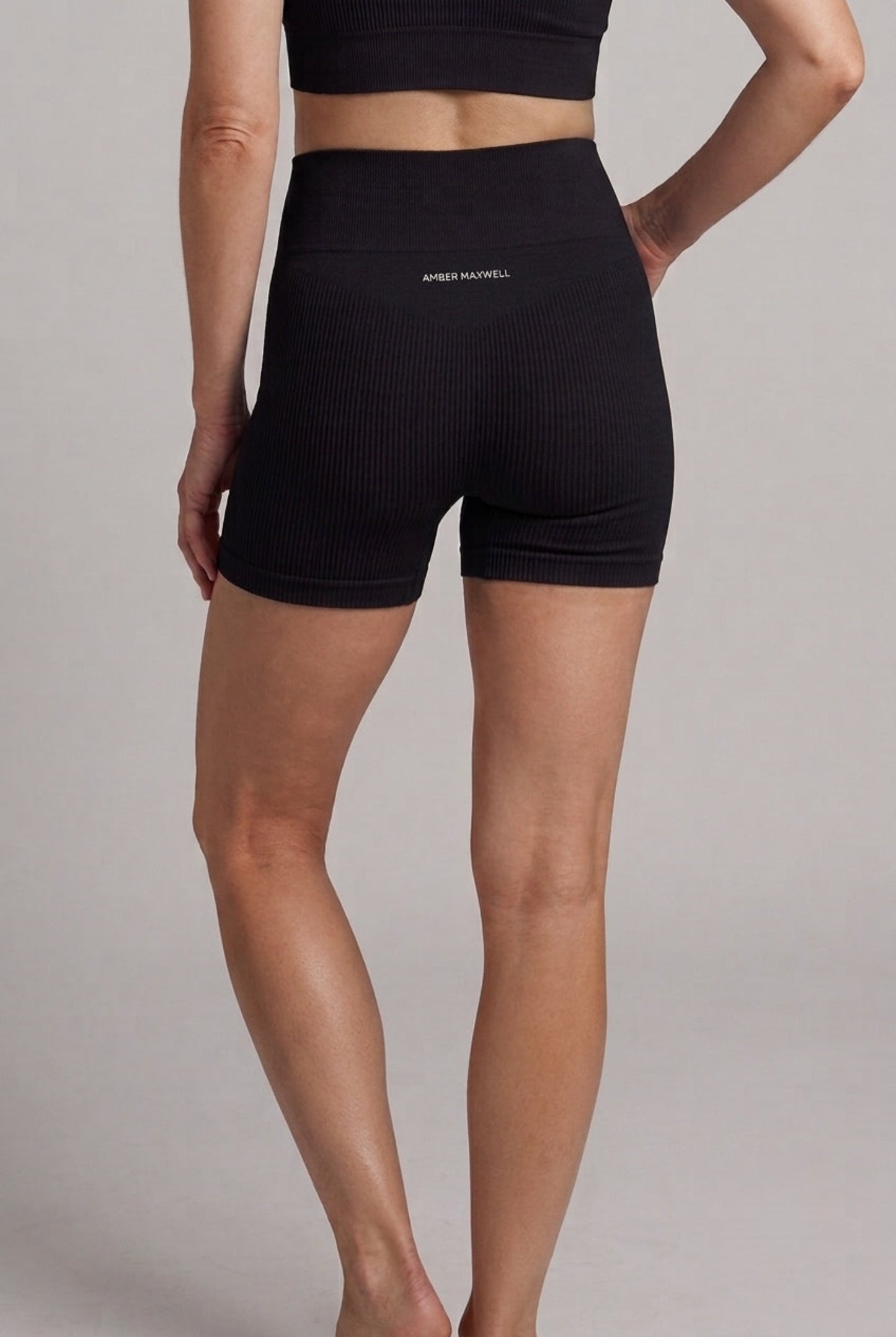 A model shown from the back wearing black ribbed high-waisted athletic shorts with a small white logo on the back waistband, against a solid light gray studio background.