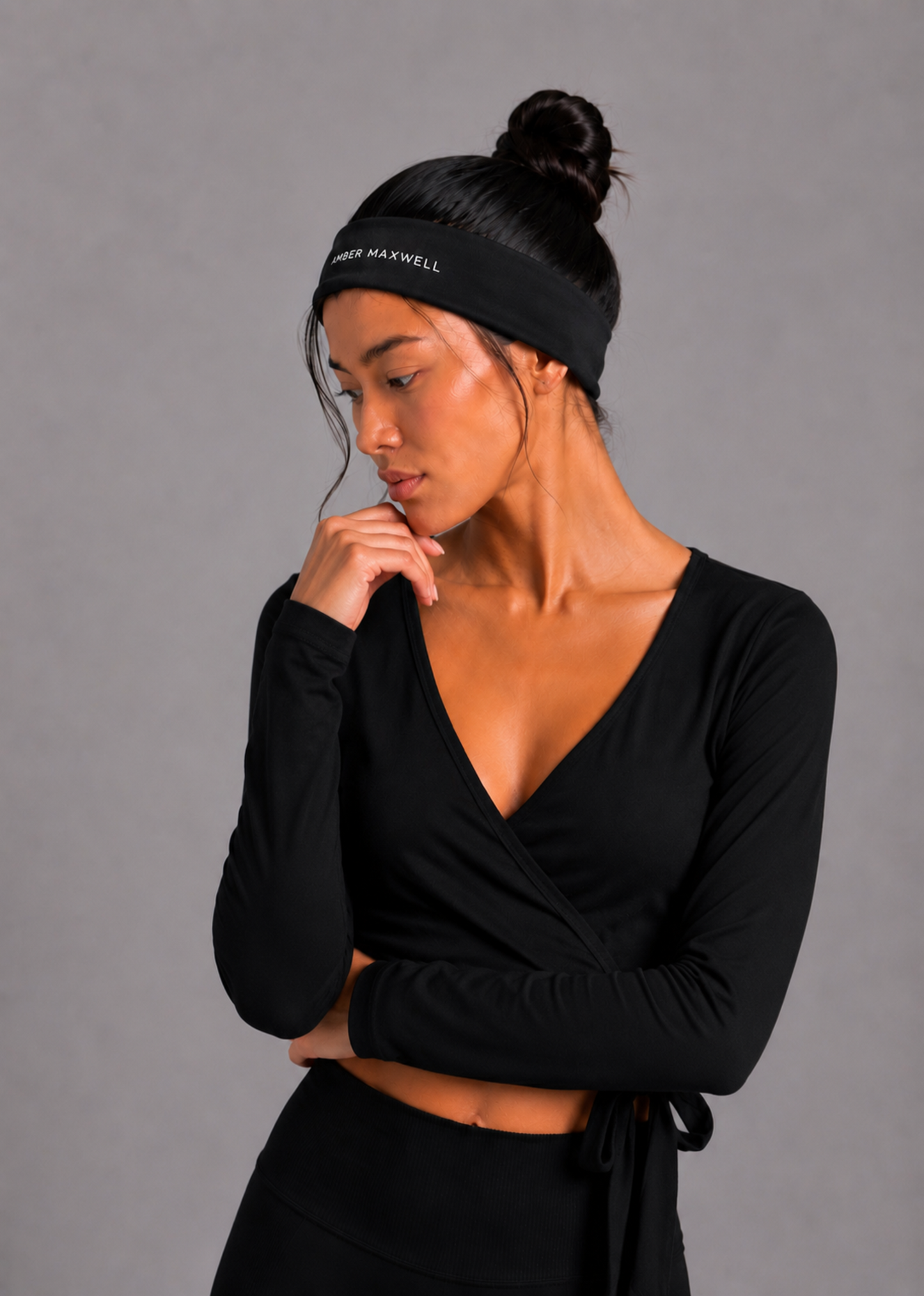 A woman wearing a black long-sleeve wrap top and matching black headband with white text reading 'Amber MAXWELL' against a solid grey studio background.