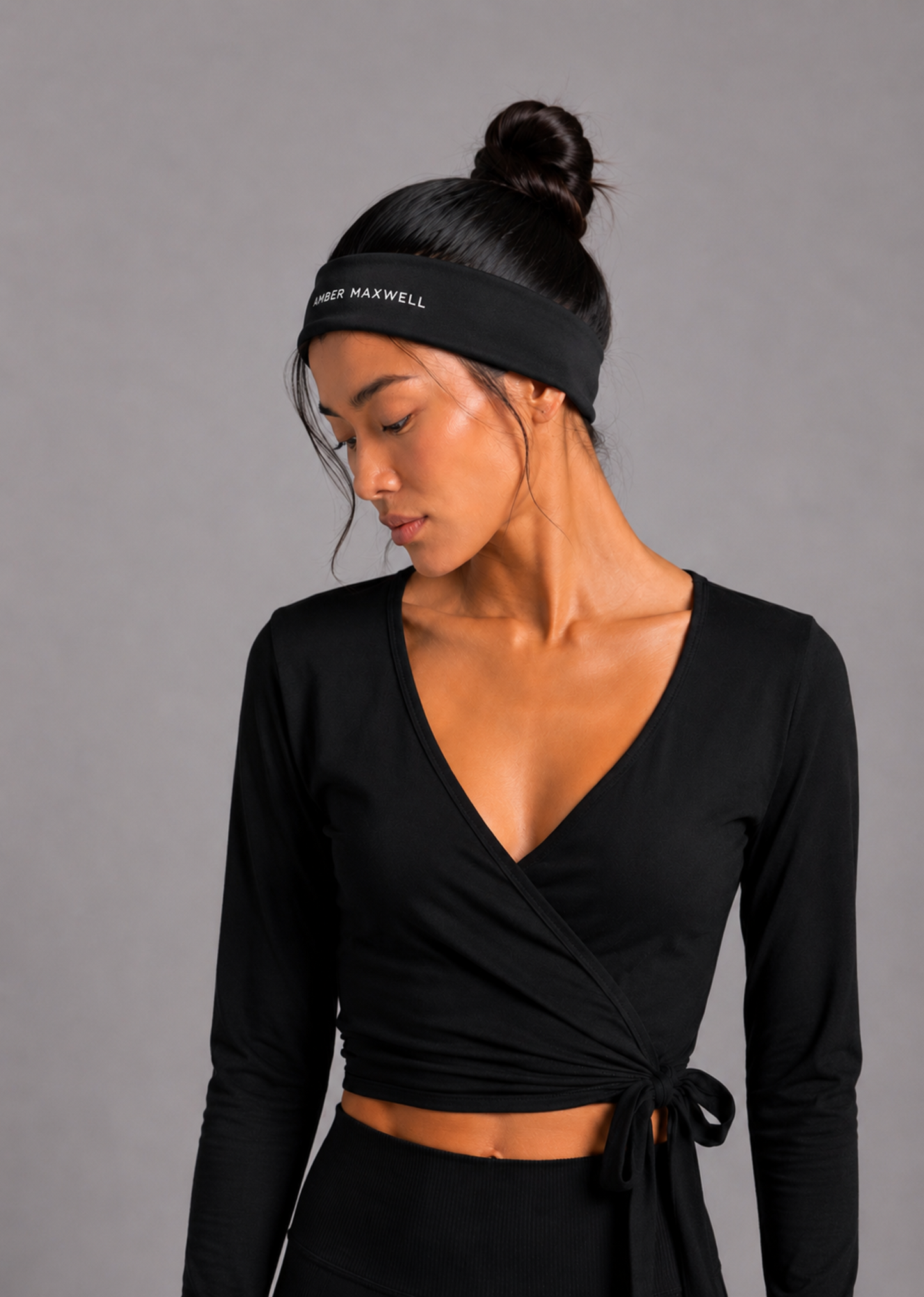 A woman wearing a black long-sleeve wrap-style crop top with a side tie, paired with a black athletic headband that reads 'Amber MAXWELL'. She is photographed against a solid gray studio background.
