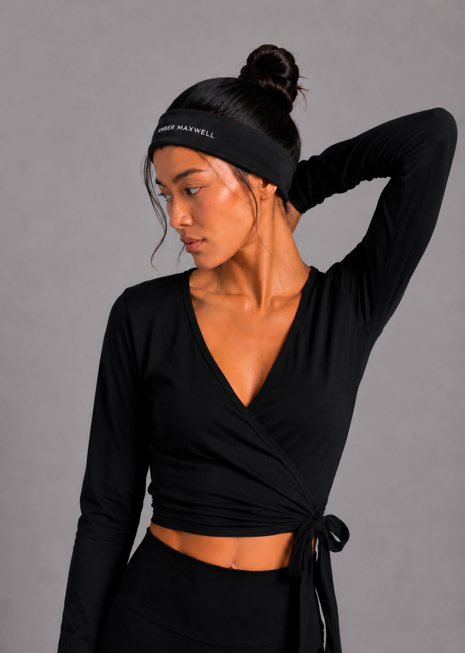 A woman wearing a black long-sleeve wrap-style crop top with a side tie, black high-waisted bottoms, and a black athletic headband with white text, posed against a solid gray studio background.