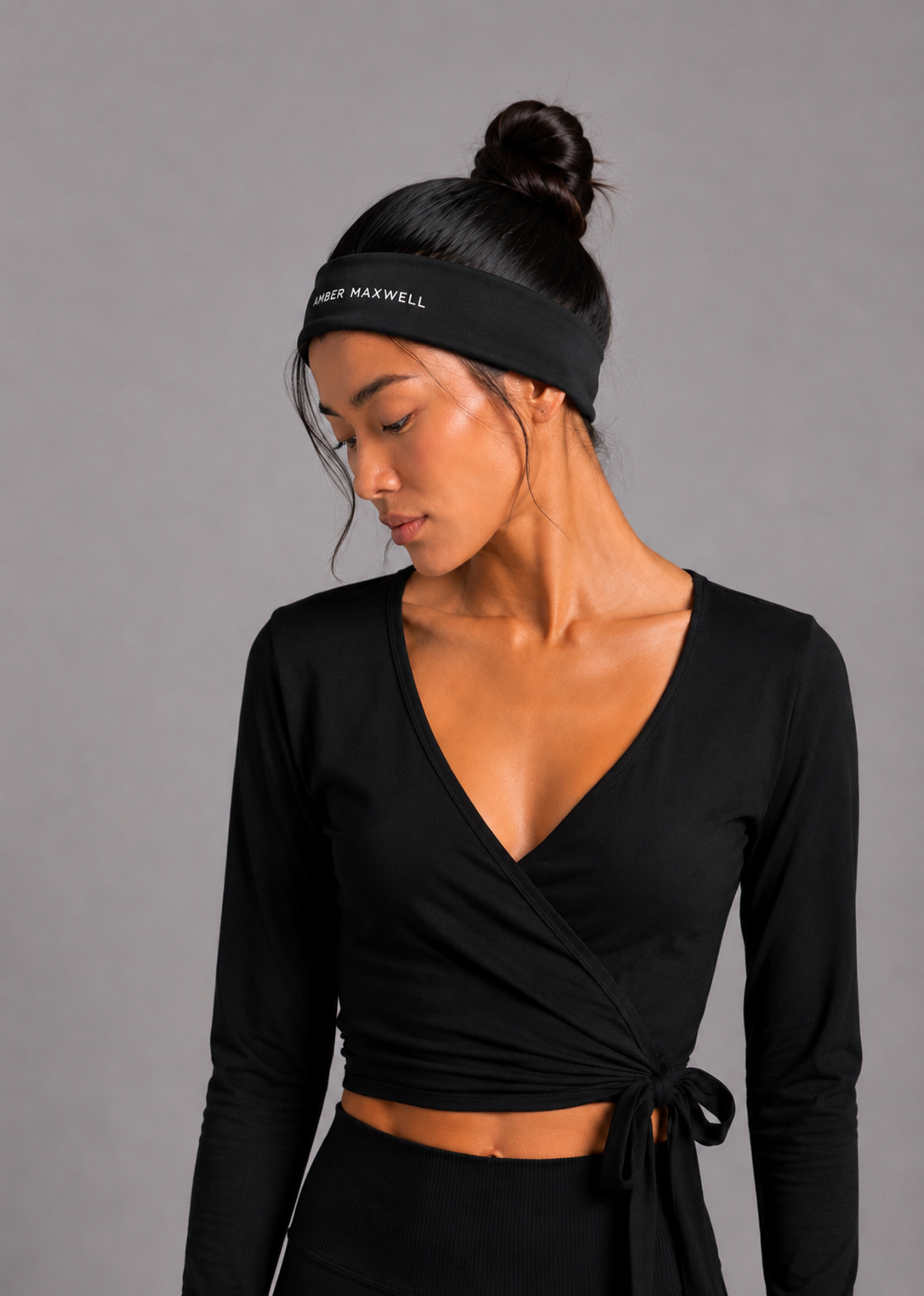 A woman wearing a black long-sleeve wrap-style crop top with a side tie, paired with a black athletic headband that reads 'Amber MAXWELL'. She is photographed against a solid grey studio background.